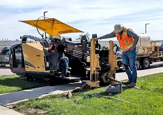 A fiber contractor in a safety vest operating a tracked directional drilling rig beside a sidewalk, with a support truck parked behind.