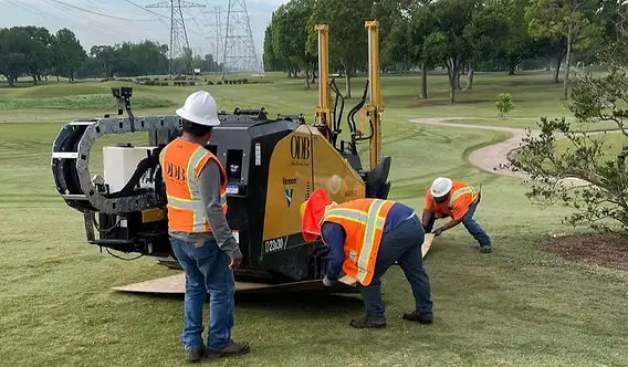 A fiber crew in safety vests positioning a tracked directional drilling rig on turf, laying protective mats beneath the machine.