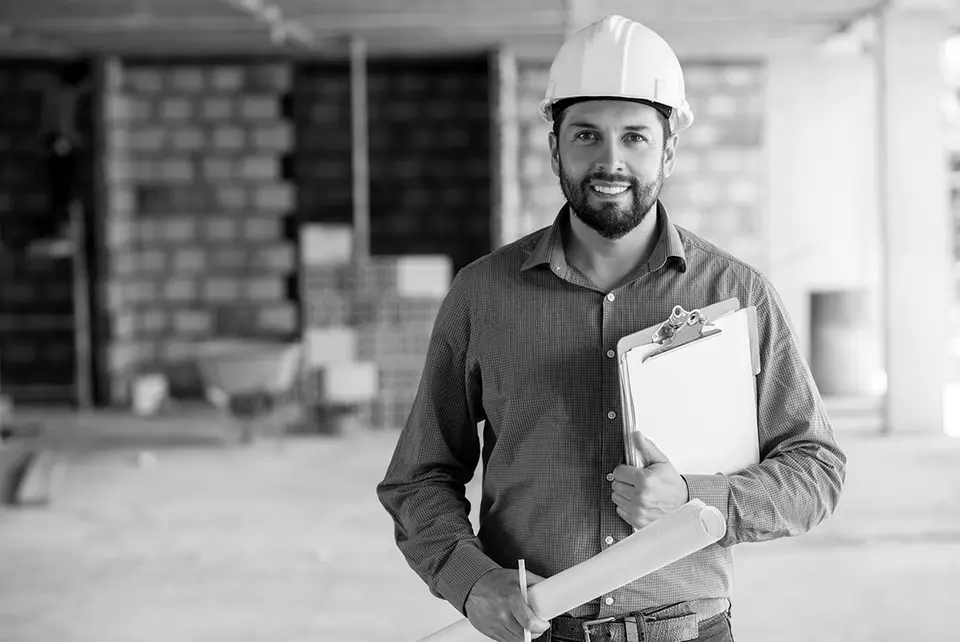 A fiber contractor in a hard hat holding a clipboard and rolled site plans at a job site.