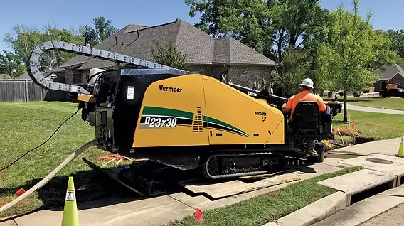A tracked horizontal directional drilling rig set up at the curb of a residential street, with a worker in an orange safety vest running the bore.