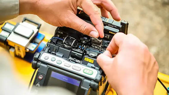 A technician’s hands using a fusion splicer to join optical fibers, with a fiber cleaver on the bench beside the machine.