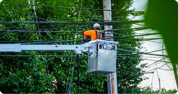 A lineworker in a hard hat and orange safety vest working from an elevated bucket lift on overhead lines amid heavy tree cover.